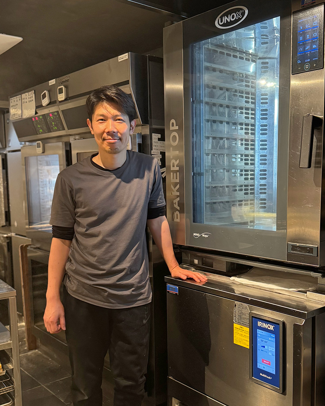 キッチンに設置されているウノックス スチームコンベクションオーブン BAKERTOPとイリノックス 加熱機能付きブラストチラー&ショックフリーザー MultiFreshの前に立っている清水宣光シェフの写真